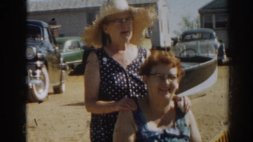 Two adult women pose together in sunlit 1963 New Jersey shore footage, with vintage cars, boats, and sandy ground behind them.