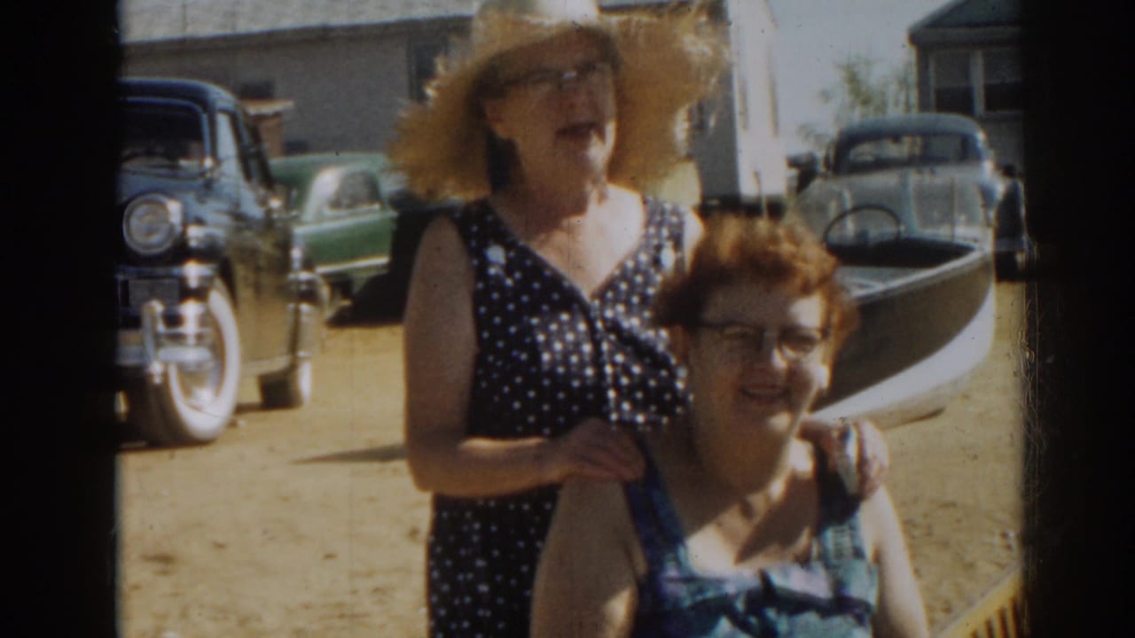 1963 New Jersey 8mm frame of two adult women smiling near a small boat and parked mid-century cars on sandy ground.