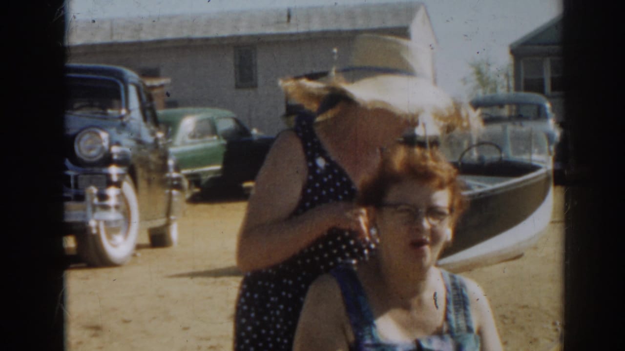 Two women near a boat and cars, with the standing woman turning under a wide straw hat.