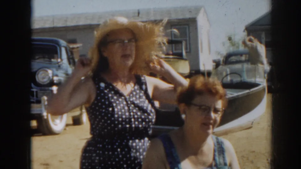 1963 New Jersey 8mm frame of a woman lifting both hands to her straw hat beside another woman in a blue swimsuit.