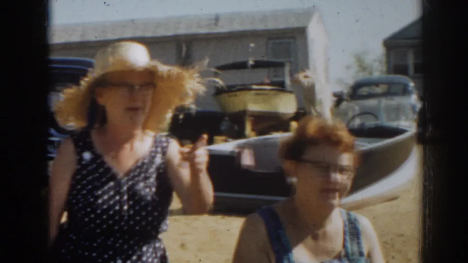 1963 New Jersey home-movie frame of two women outdoors, one gesturing in a straw hat near a boat and parked cars.