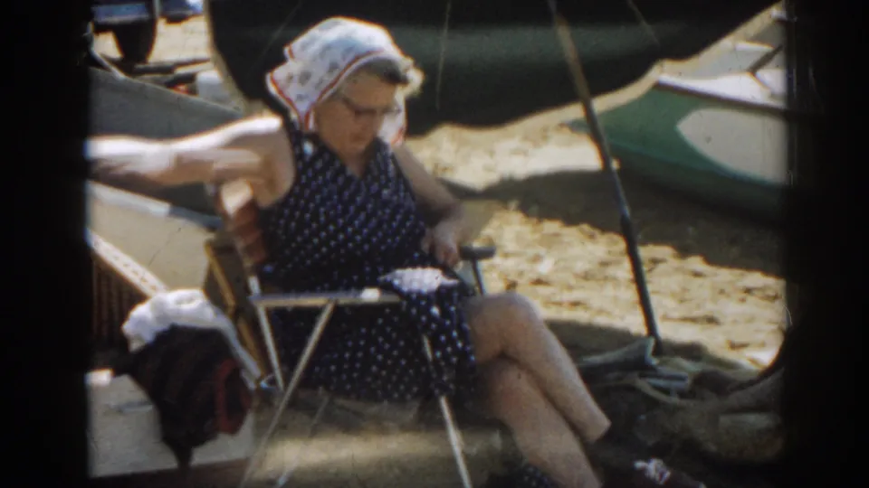 Adult woman wearing glasses and a headscarf looks down from a folding chair on a sandy New Jersey beach in 1963.