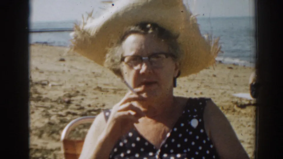 Close home-movie view of an adult woman in glasses and a large straw hat on a New Jersey beach in 1963.