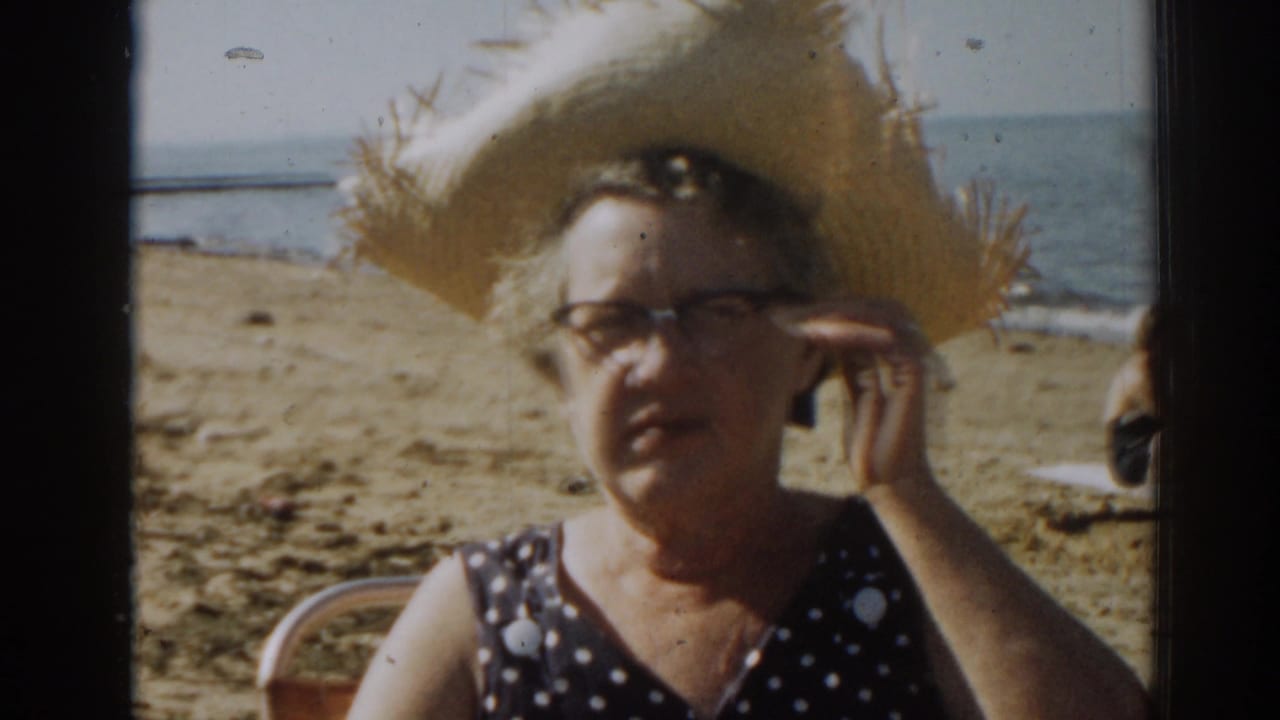 Woman in straw hat and glasses sits on a sandy beach near the ocean.