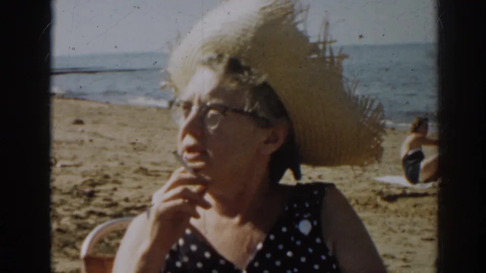 The woman turns away from the camera while holding a cigarette, with sand, water, and another beachgoer visible behind her.
