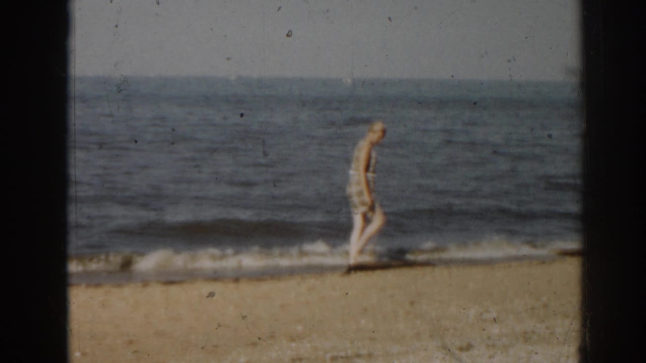 Woman in a white skirt walks near small waves on a sandy beach.