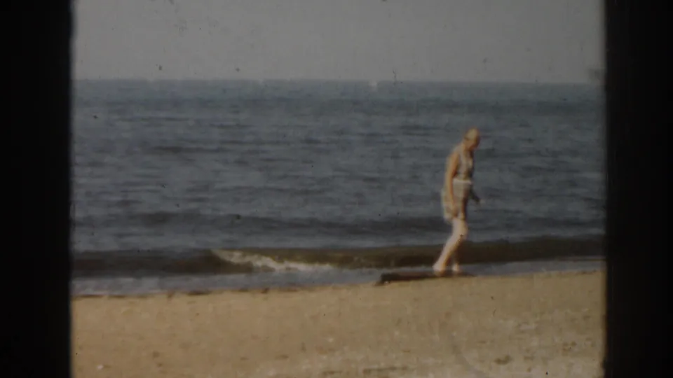 1963 New Jersey beach frame with a woman in light clothing walking parallel to the surf on 8mm home-movie film.