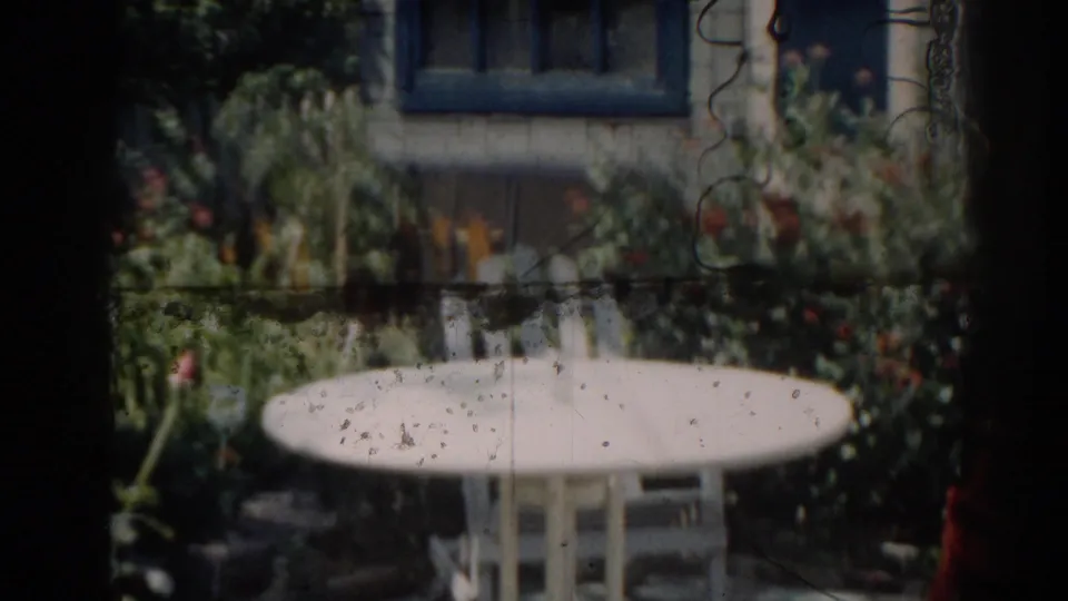 Backyard garden view with a white round table, chairs, flowering plants, and a blue-trimmed window in 1963 home-movie footage.