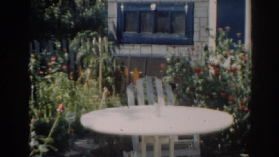 1963 home-movie frame of a backyard garden with white patio furniture, dense plants, flowers, and a blue-framed house window.