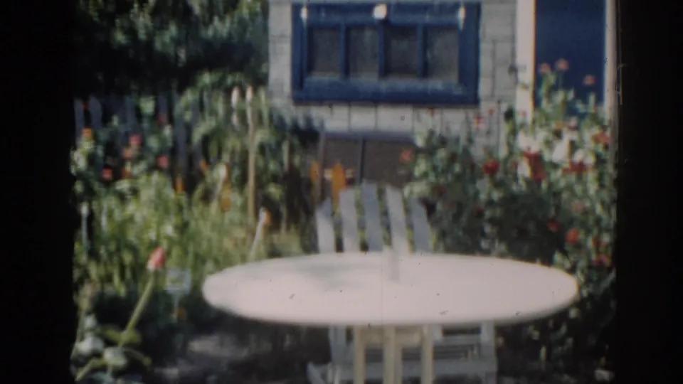 1963 New Jersey home-movie frame of a backyard seating area framed by flowers and a house with blue window trim.