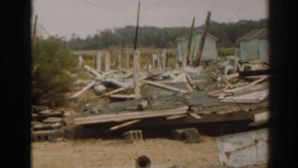 1963 New Jersey 8mm frame showing a damaged or destroyed home site with scattered wood, posts, rubble, and nearby structures.