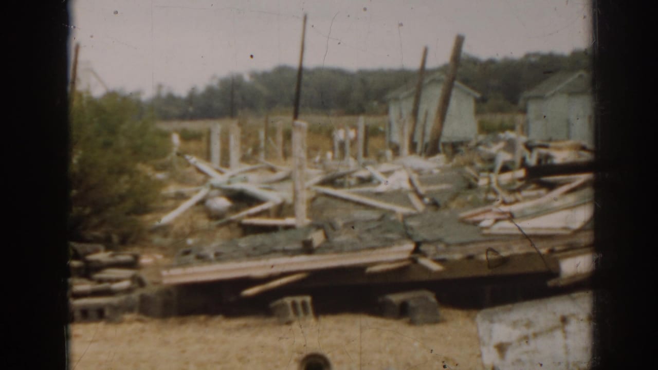 Collapsed lumber and scattered debris near small houses, seen through a dark vertical frame.