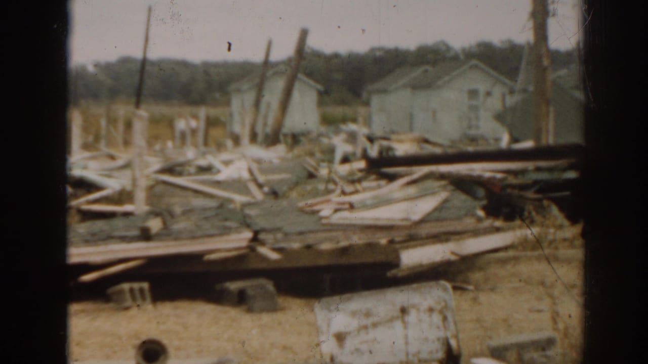 Wrecked wooden debris lies across the ground with small houses and poles in the background.