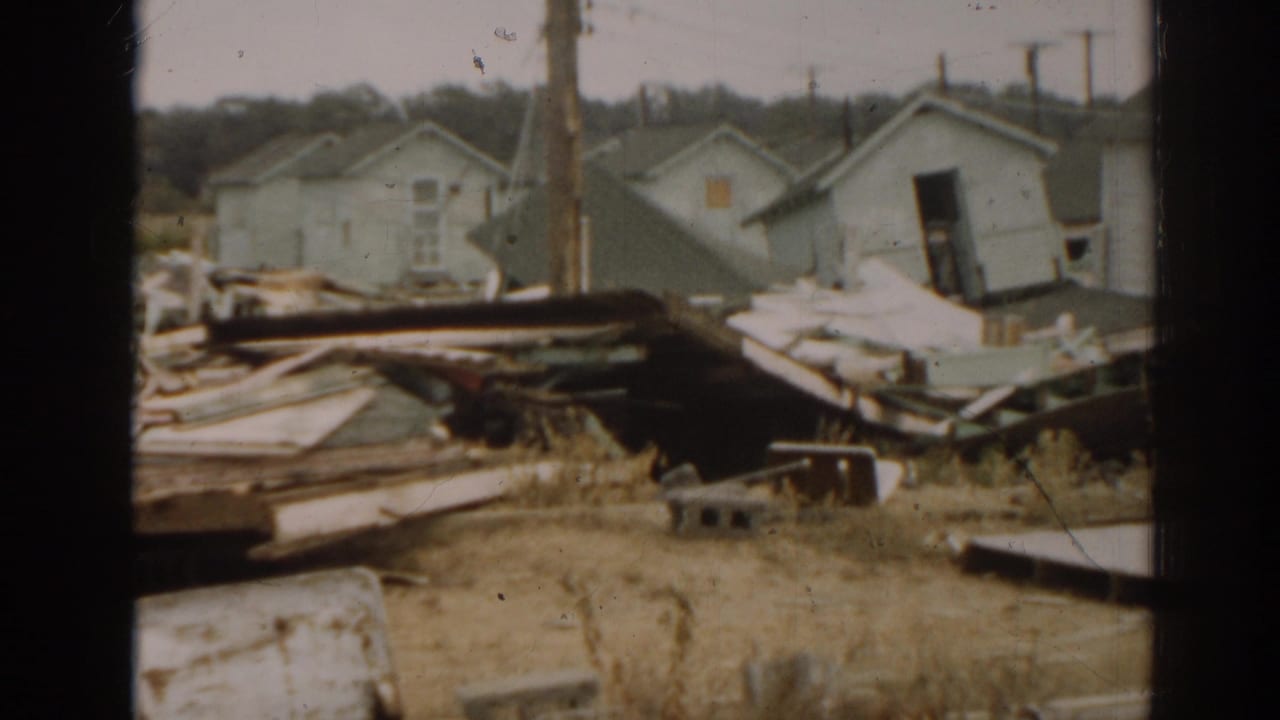 Debris-strewn ground with small light-colored buildings and a leaning damaged structure.