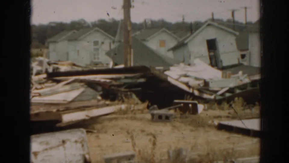 1963 New Jersey home-movie frame with a leaning small structure, pale buildings, wood debris, cinder block, and utility poles in view.