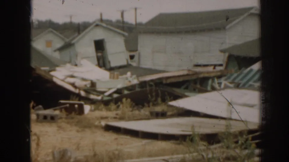 1963 New Jersey frame showing a tilted small building, scattered boards, dry vegetation, and larger pale structure in the background.