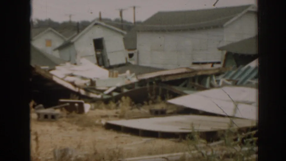 1963 New Jersey 8mm frame with a damaged pale structure, broad loose panels, dry grass, and a larger building behind it.