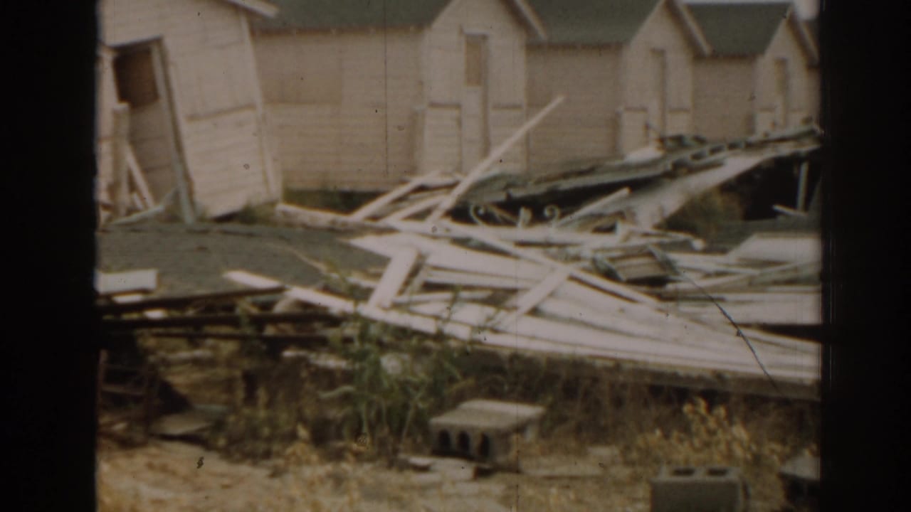 Damaged tan wooden shacks with collapsed boards and debris in a yard.