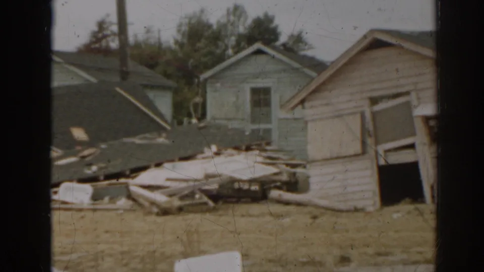 1963 New Jersey frame showing a damaged wooden shed, nearby rooftops, scattered boards, and dry grass in a storm aftermath scene.