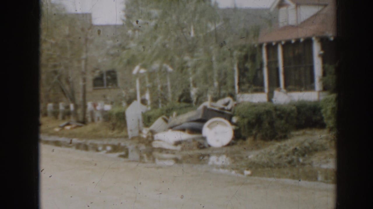 Residential yard with porch, shrubs, discarded items, and a vintage car partly visible.