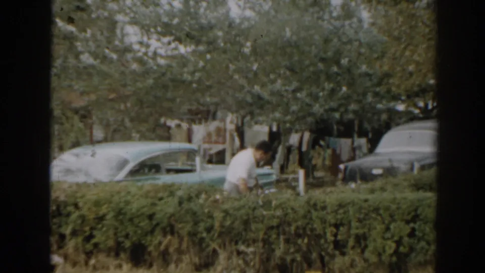 1963 New Jersey home-movie frame showing a man working by a hedge, with vintage cars and a clothesline in the yard.