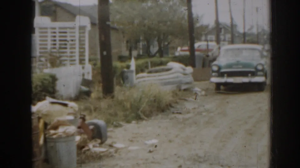 1963 New Jersey home-movie view down a residential driveway with a dark vintage car, trash cans, hedges, and utility poles.