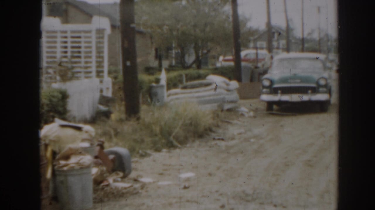 Classic sedan parked on a dirt driveway beside a cluttered yard and trash cans.