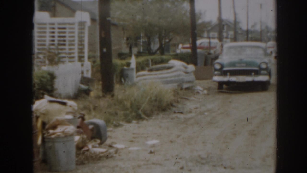 Parked sedan faces the camera from a dirt driveway with clutter along the yard edge.