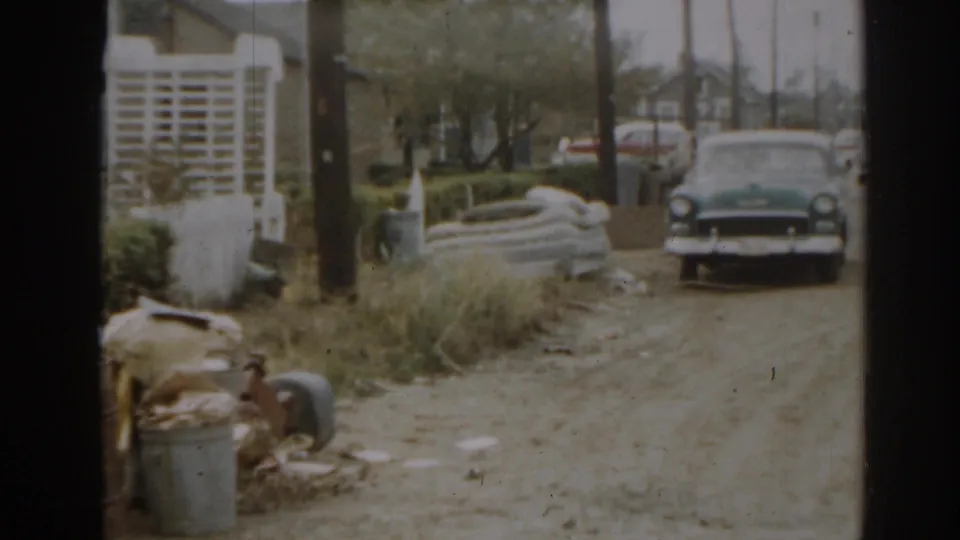 1963 New Jersey residential view with a parked sedan, lattice fence, trash can, overgrown grass, and household debris near the driveway.