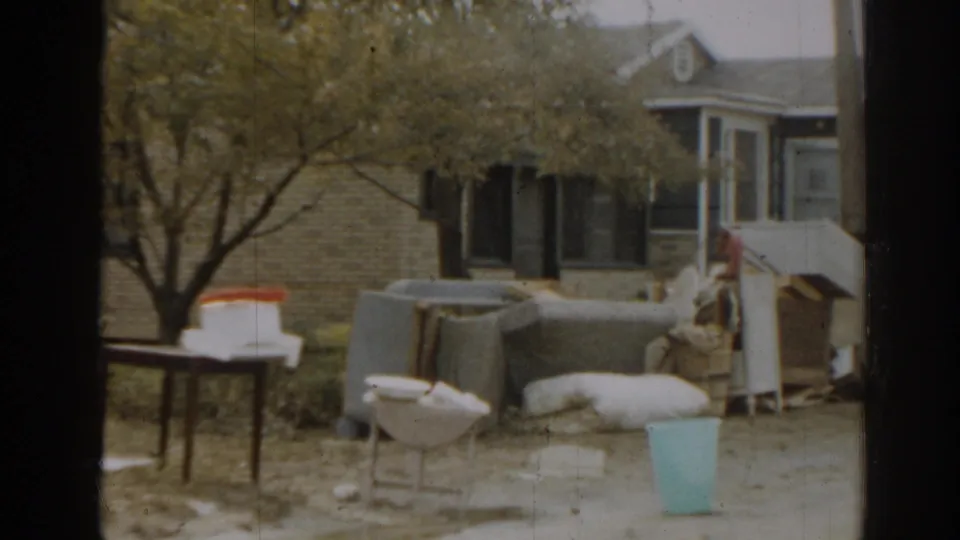 1963 New Jersey home-movie frame of a suburban yard with furniture, a table, a blue bucket, trees, and house fronts.