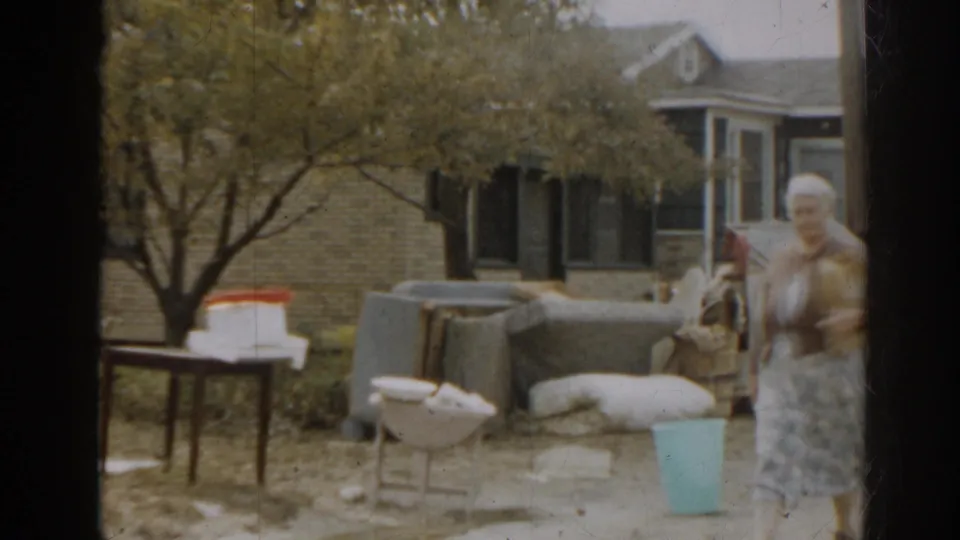 1963 New Jersey home-movie frame showing a woman walking near yard furniture, a blue bucket, trees, and residential houses.