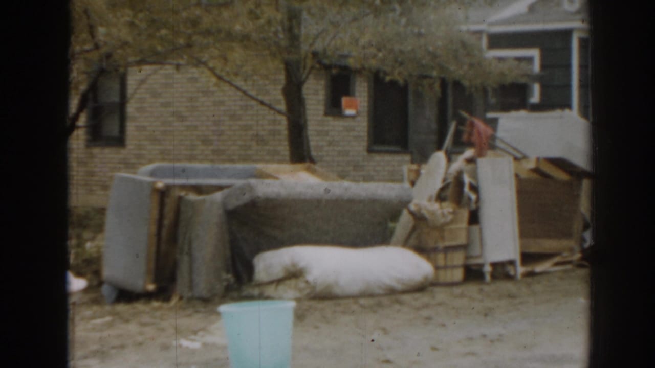 Outdoor pile of furniture and bedding beside a brick house and tree, with a pale bin in front.