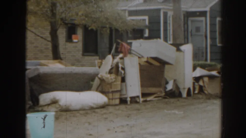 1963 New Jersey home-movie frame showing stacked furniture, bedding, cardboard, and neighboring house fronts in a residential setting.