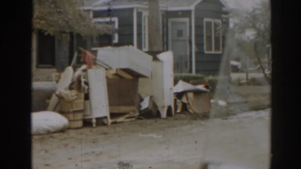 1963 New Jersey home-movie frame showing a residential exterior, curbside furniture debris, bedding, cardboard, a pole, and the edge of a street.