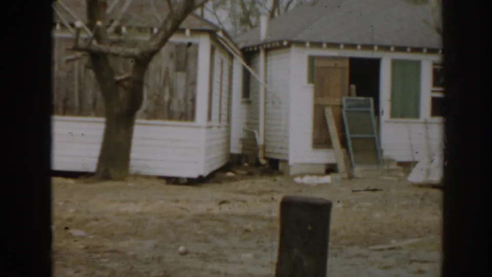 1963 New Jersey home-movie frame showing a small white outbuilding, open doorway, leaning board, and bare yard.
