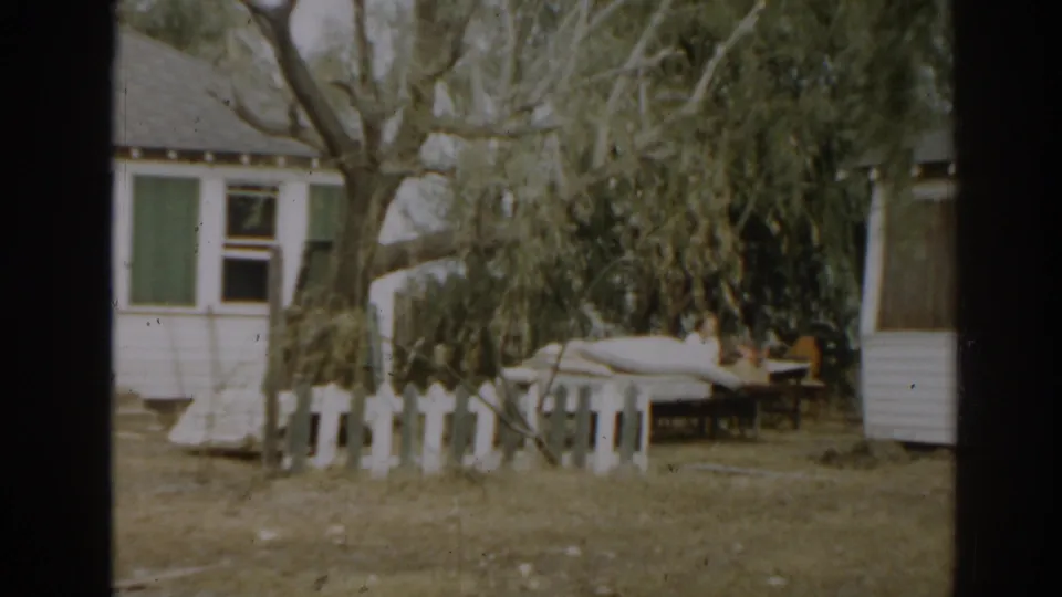 1963 New Jersey frame showing a residential yard with a white fence, overgrown branches, debris, and part of a small building.