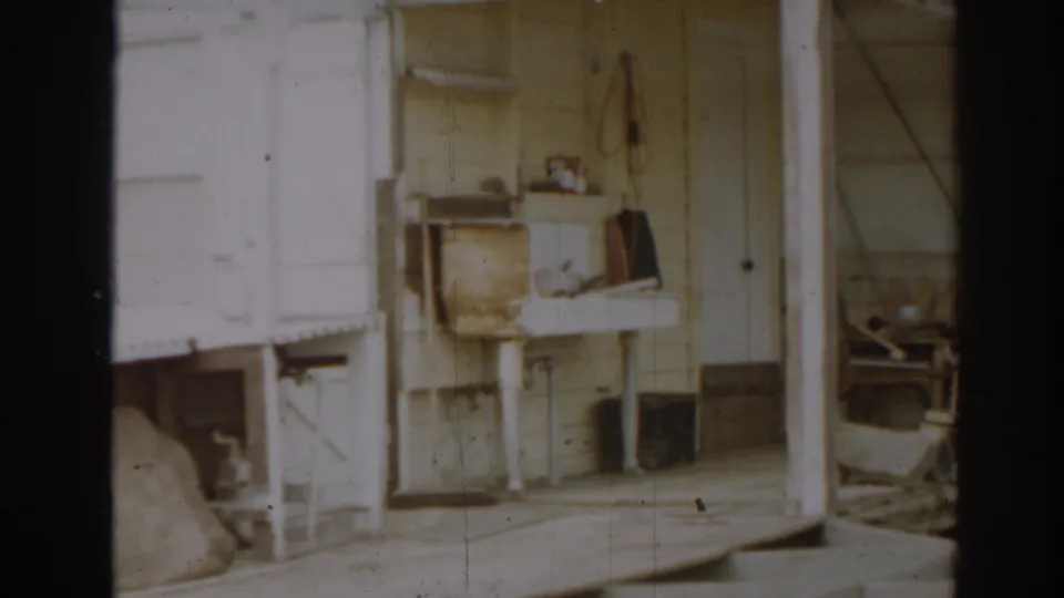 1963 New Jersey home-movie view of a farm porch with a utility sink, shelves, tools, pale siding, and shadowed openings.