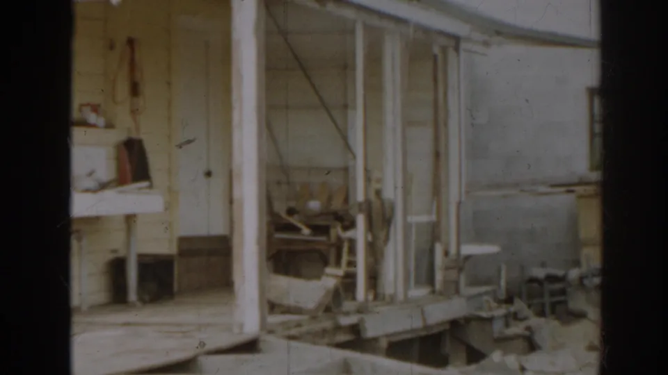 1963 New Jersey farm outbuilding corner with porch posts, pale siding, a low work surface, scattered tools, and yard space beyond.