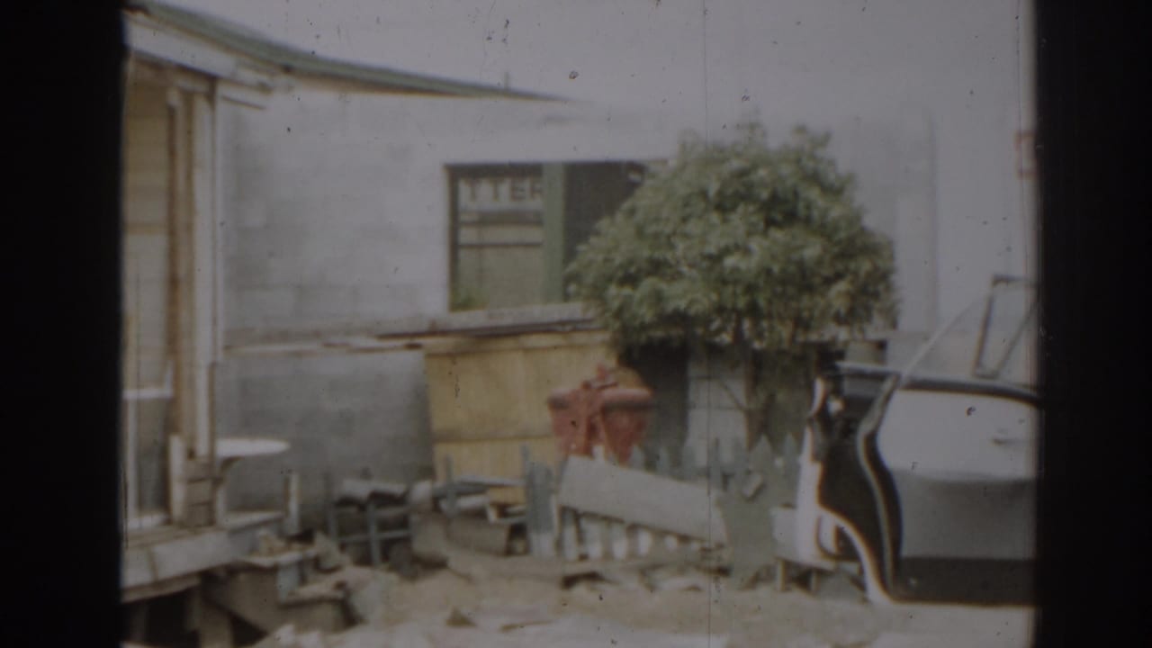 House exterior with porch damage, shrub, fence rails, and the edge of a car door.
