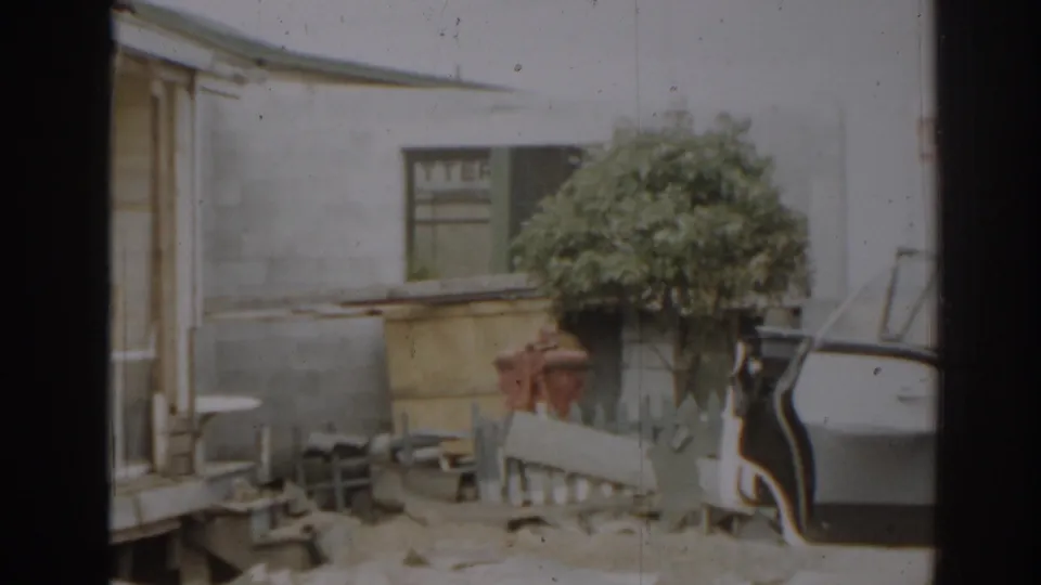 1963 New Jersey frame showing a damaged residential exterior with porch debris, a shrub, fence rails, and a car door edge.