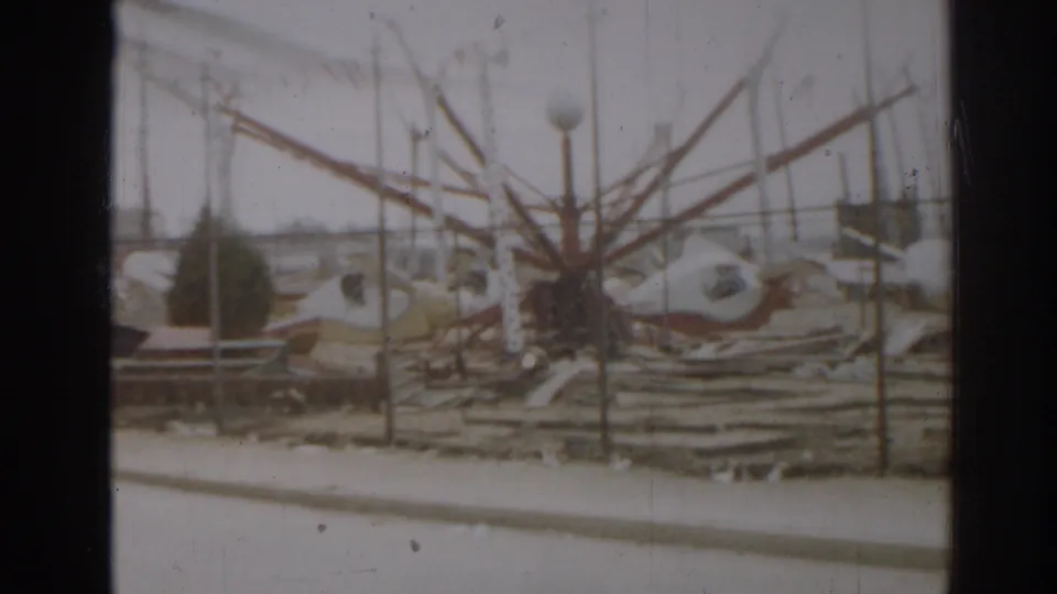 1963 New Jersey home-movie frame showing a damaged amusement-ride-like structure behind fence rails with scattered debris.