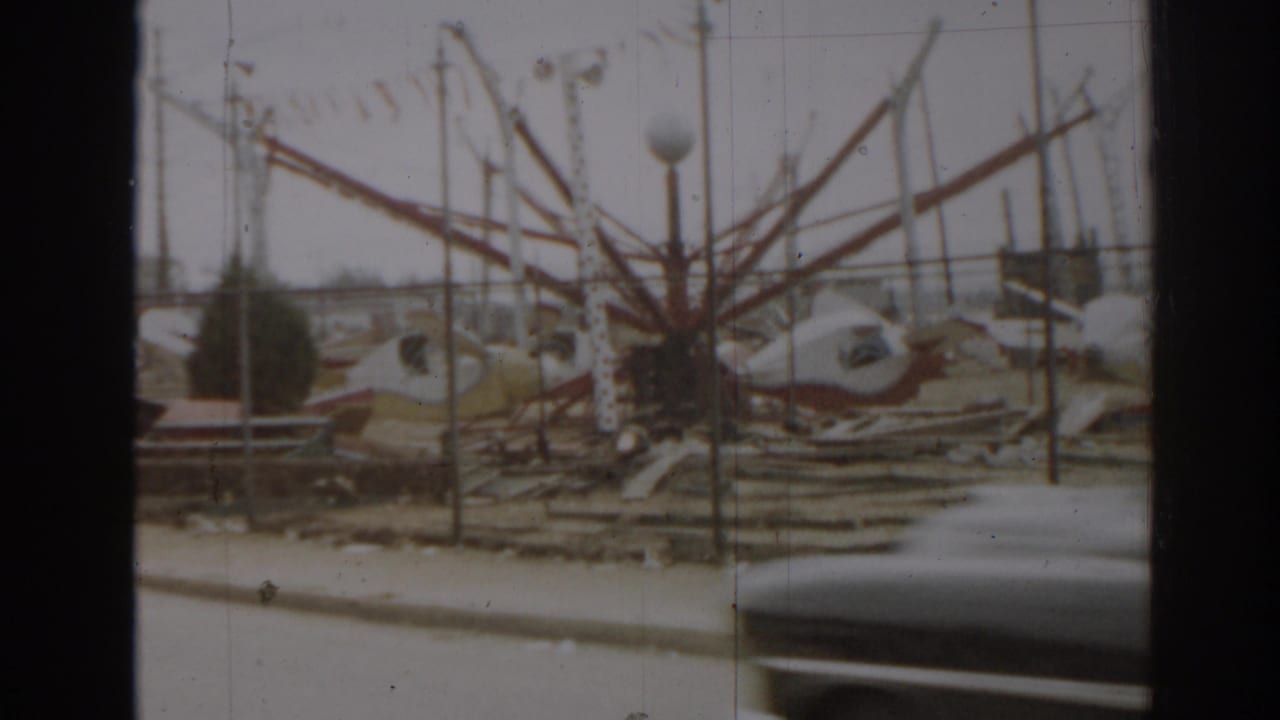 Damaged amusement ride structures behind a chain-link fence as a dark car passes in the foreground.
