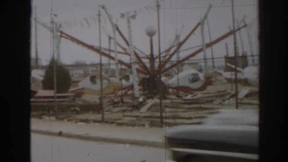 1963 New Jersey 8mm frame showing wrecked amusement-park ride frames and debris behind roadside fencing, partly blocked by a passing car.