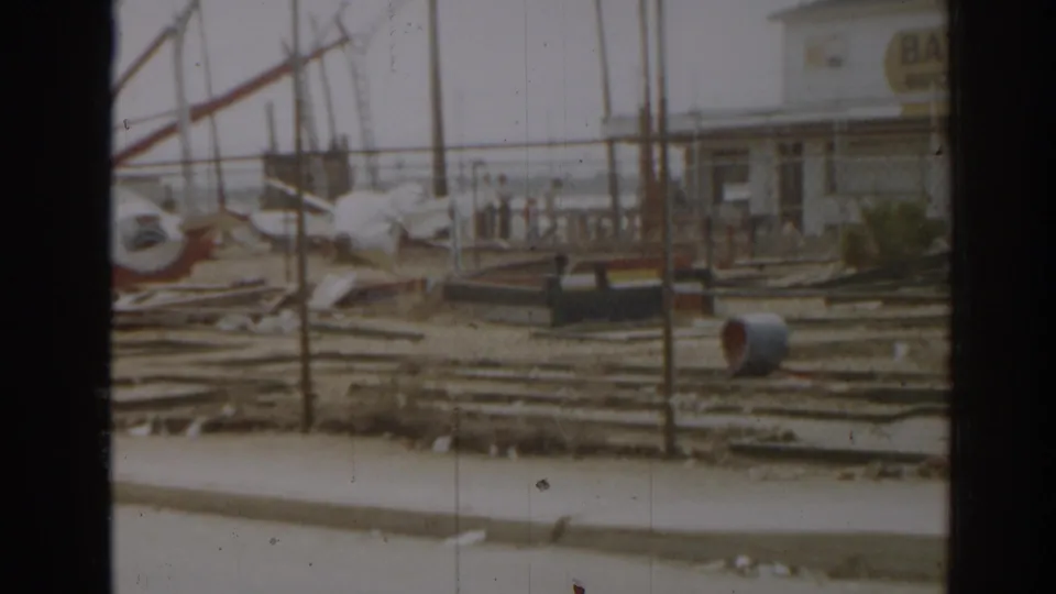 1963 New Jersey home-movie frame showing ruined amusement-park equipment, debris, fencing, and a roadside curb from a moving vehicle.