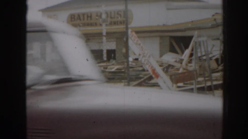 Passing car partly obscures a damaged bath house area with scattered debris, fencing, and a visible warning sign in 1963 New Jersey footage.