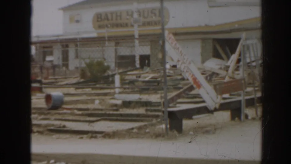 A New Jersey 1963 home-movie view of a bath house facade with debris, railings, and leaning damaged elements.
