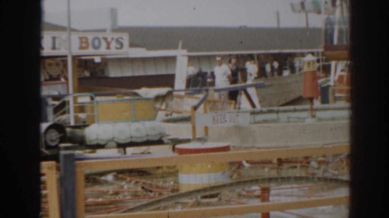 1963 New Jersey home-movie frame showing a storm-damaged amusement park ride area with signage, railings, debris, and onlookers.