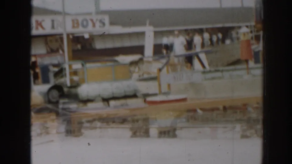 Soft 8mm frame of New Jersey amusement park storm damage, showing a ride area, BOYS signage, debris, railings, and distant onlookers.