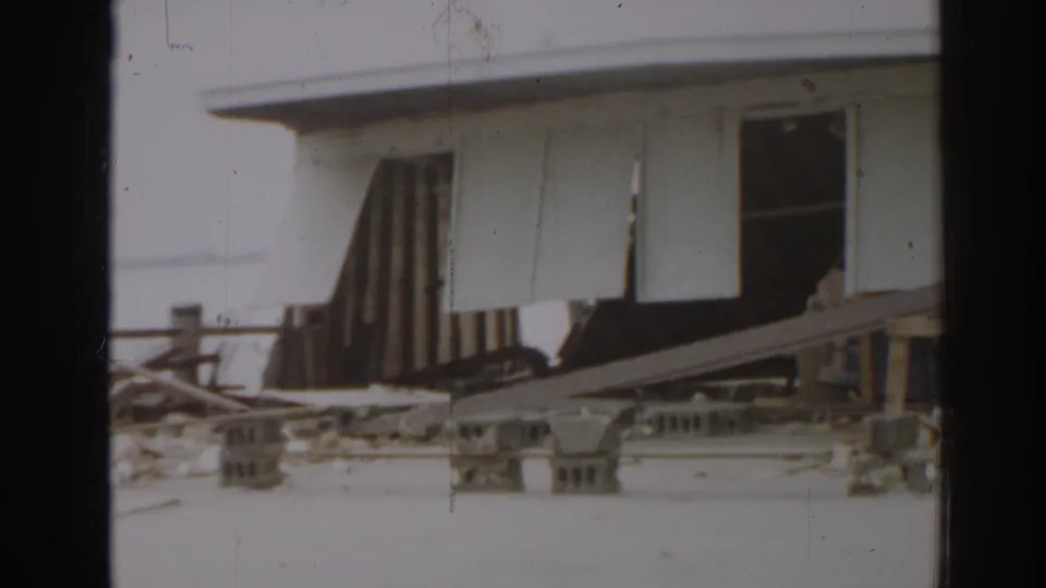 1963 New Jersey home-movie frame showing a damaged white amusement-park building with torn siding or roofing and debris below.