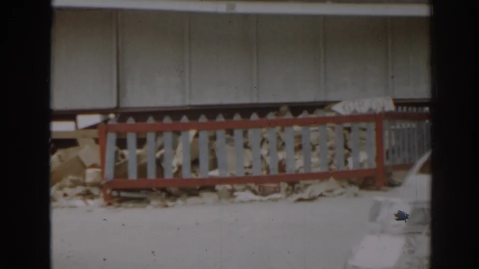 A red and white fence stands before a damaged building with debris visible behind it in this 1963 New Jersey home-movie frame.